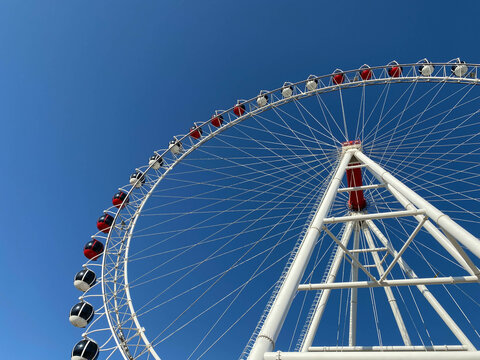 A New Very Tall White Ferris Wheel With Comfortable Booths For People Is Slowly Rotating In A Circle In The Center Of A Big City Against A Clear Blue Sky