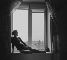 Beautiful European girl sits on the window in a black bodysuit