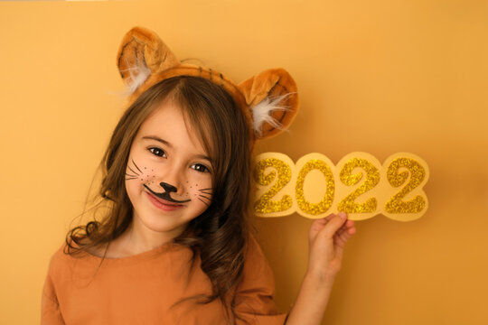 Cute curly-haired brunette girl with tiger aquagrim holds golden shining numbers 2022 in her hand. A headdress with tiger ears. New Year. Child in a tiger costume smiles sweetly and looks at camera