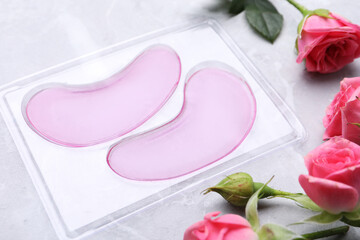 Package of under eye patches and rose flowers on light grey table. Cosmetic product