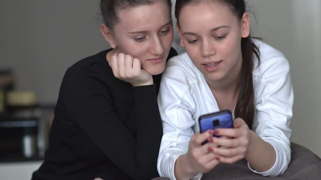 two young women sit on couch at home looking at smartphone screen working reading or watching news about covid 19 coronavirus chatting in social media surfs the inernet