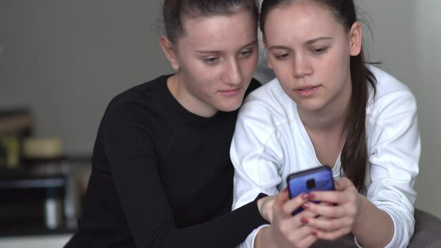 two young women sit on couch at home looking at smartphone screen working reading or watching news about covid 19 coronavirus chatting in social media surfs the inernet