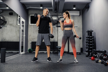 Fototapeta na wymiar Fitness couple doing arm exercises together and lifting dumbbells indoor modern gym. Front view of a couple dressed in sportswear doing arm exercises and they make eye contact to support each other