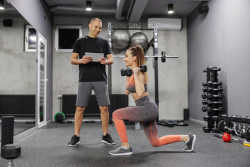 Fototapeta na wymiar Measuring the success of fitness training Woman in sportswear makes a step forward in the gym and holds dumbbells in her hands while a coach observes her progress and enters data into a digital tablet