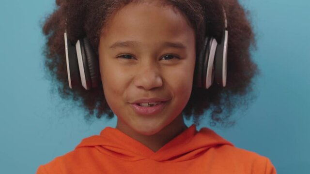 Cute African American girl enjoys singing song and listening the music in headphones on blue background in slow motion. Close up of kid talking looking at camera.