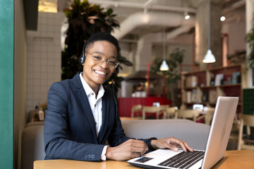 Beautiful african american woman looking at camera and smiling, working in coworking cafe, video communication using headset business woman freelancer