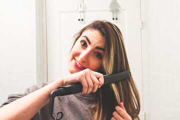 Mirror point of view of a caucasian young woman straightening her hair with a hair straightener in...