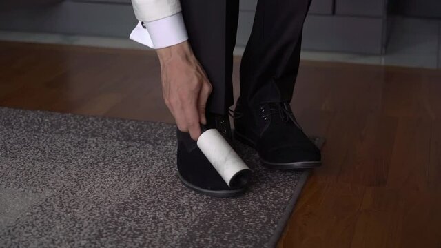 Man in a classic suit gets dressed, cleans shoes and clothes with adhesive tape from wool and pellets. Groom or businessman wears for holiday or work.Man in white shirt and black pants. Sticky roller.