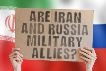 The question " Are Iran and Russia military allies? " on a banner in men's hand with blurred Russian and Iranian flag on the background. Support. Weapon. Tehran
