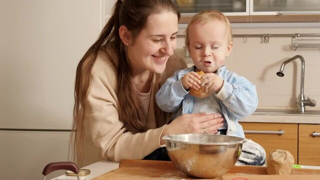 Cute smiling baby boy with mother eating fresh bread and playing with flour. Concept of little chef, children cooking food, healthy nutrition.