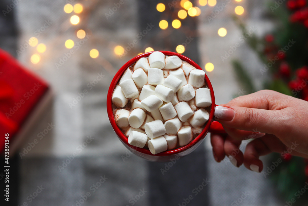 Poster woman's hand holding a cup of hot chocolate with marshmallows on blurred holiday background. winter 