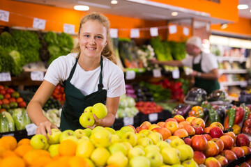 Fifteen-year-old girl who works part-time in a store as a trainee saleswoman puts fresh apples on...