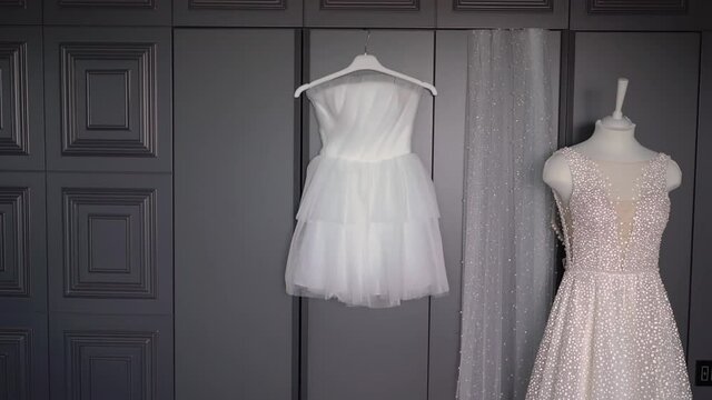 Two wedding dresses, white bridal gown hanging on hanger in bedroom. Morning of the bride. Long and short, white and pink. Bride veil in store.