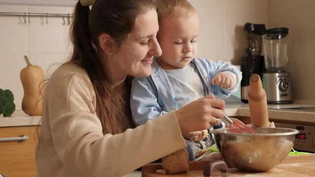 Smiling young mother with little baby son making bread dough in bowl on kitchen. Concept of little chef, children cooking food, healthy nutrition.