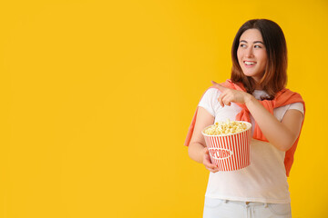 Young Asian woman with tasty popcorn pointing at something on yellow background