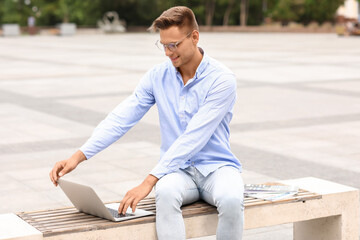 Fototapeta na wymiar Handsome young businessman using laptop on bench outdoors