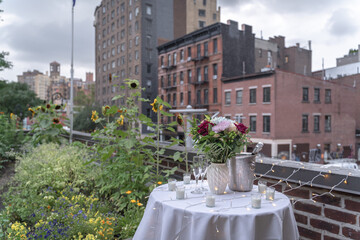 A bouquet of flowers sits on top of a table draped with white tablecloth, candles and fairy lights in celebration of a surprise proposal. Buildings in West Villages are in the background.