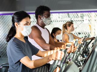 Portrait of young adult woman training on elliptical cross trainer in gym, all people wearing face...