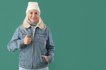 Beautiful mature woman in warm clothes showing thumb-up on color background