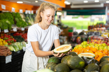 Young positive girl choosing sweet fresh melon at grocery section of supermarket