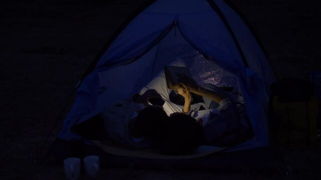 a guy and a girl lie in a tent at night reading a book on which they shine a flashlight