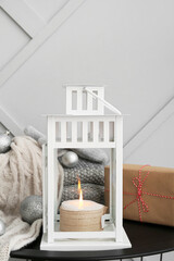 White lantern with burning candle, gift, Christmas balls and warm clothes on table near light wall