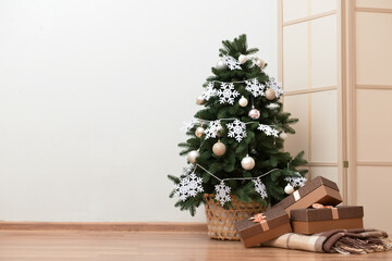 Beautiful Christmas tree in basket decorated with snowflakes, balls and gift boxes near light wall