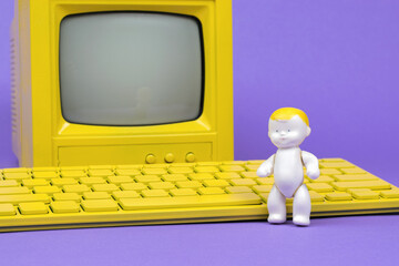 A baby figurine near a yellow keyboard and a yellow monitor on a purple background.