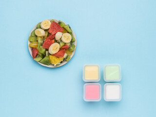 Four cups of yogurt and a bowl of fruit salad on a blue background. Flat lay.