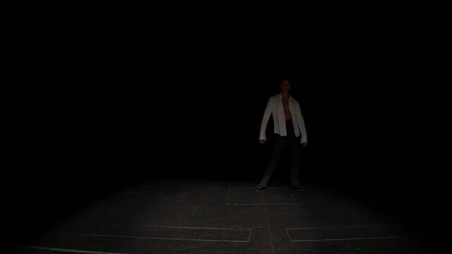 Ballroom dancer comes out of darkness onto marked stage with slow,flexible steps forward and sideways in Latin American tango dance style,hand rotation,swing,lunges to sides.Solo performance element.