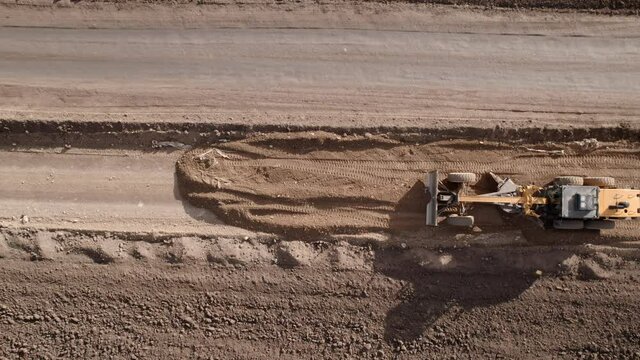Aerial drone shot of road grader smoothing gravel surface for new road. Scraper leveling ground, working at construction. Earthmoving, excavations, digging on soils.