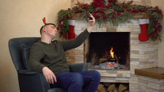 A young man sits in an armchair by the fireplace with antlers on his head and takes a selfie. Christmas mood.