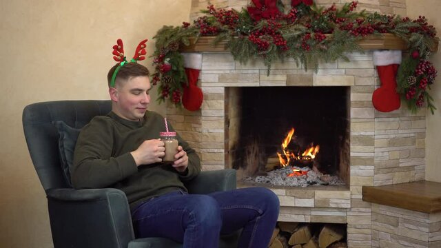 A young man sits in an armchair by the fireplace with horns on his head and drinks cocoa. Christmas mood.