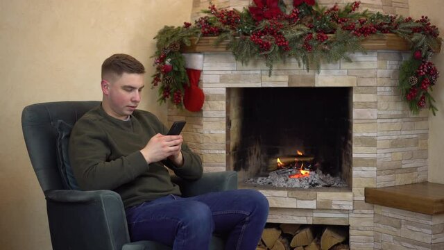 A young man is sitting in an armchair by the fireplace and texting in a smartphone.