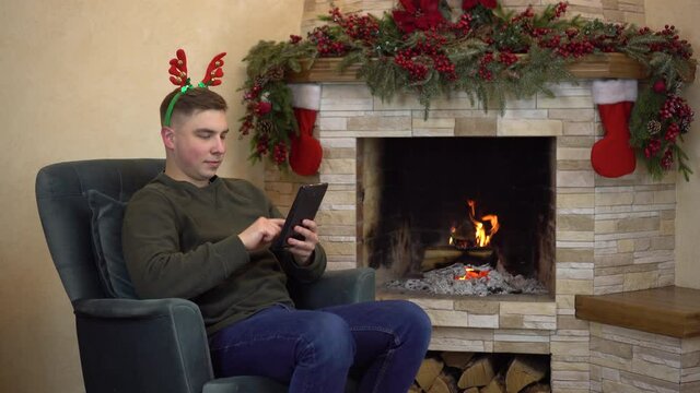 A young man is sitting in a chair with horns on his head by the fireplace and texting from a tablet.