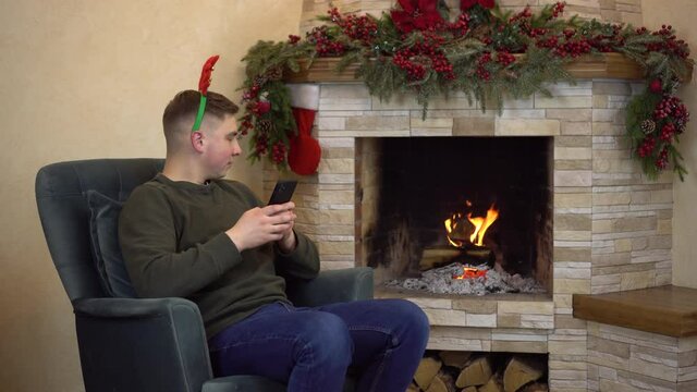 A young man sits in an armchair by the fireplace with horns on his head and writes in a smartphone.