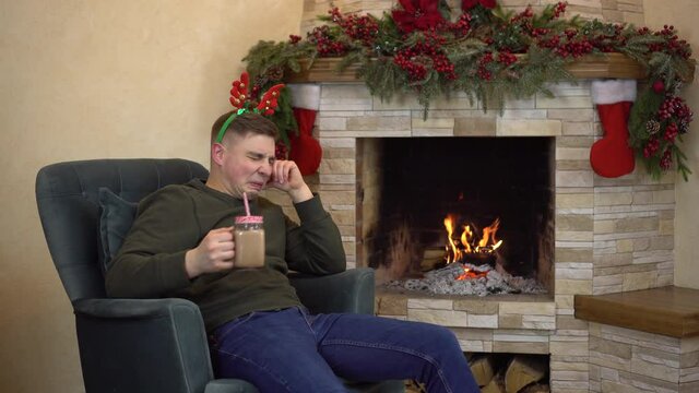 A sad young man sits in an armchair by the fireplace with horns on his head and drinks cocoa and cries. Sad Christmas.