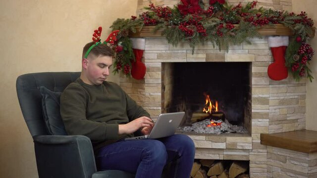 A young man sits in an armchair by the fireplace with horns on his head and is typing on a laptop. A man writes an email to Santa Claus.