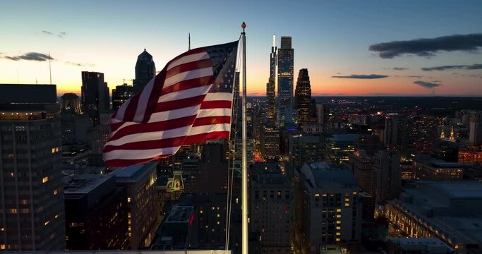 Slow motion of American flag waving at sunset against skyscraper highrise cityscape at night. USA pride in metropolitan city.
