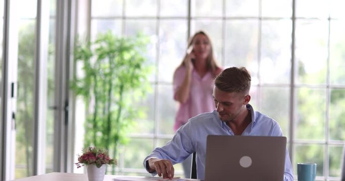 Caucasian husband working online with laptop computer and drinking coffee at work desk while wife walk around have loud noise funny gossip conversation on phone be considerate move to other room.