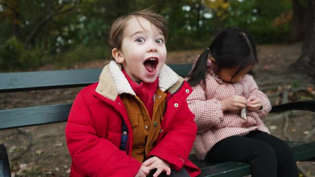 View of a little boy looking at the camera and making funny faces while his friend busy unwrapping a chocolate bar in a park on a wintry evening.