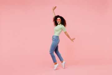Full size body length young curly latin woman 20s wears casual clothes sunglasses stand on toes dance lean back have fun spreading hands isolated on plain pastel light pink background studio portrait.