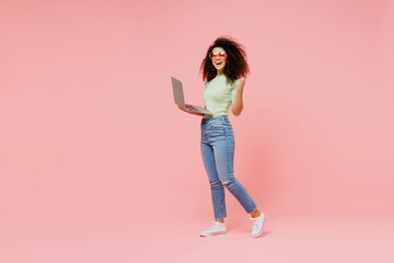 Fototapeta na wymiar Full size body length fun young curly latin woman 20s wears casual clothes go walk hold use work on laptop pc computer do winner gesture isolated on plain pastel light pink background studio portrait.