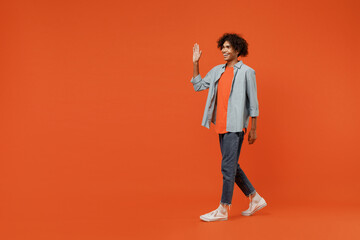 Full body side view young smiling black student man 50s wearing blue shirt t-shirt waving hand walk go strolling isolated on plain orange color background studio portrait. People lifestyle concept