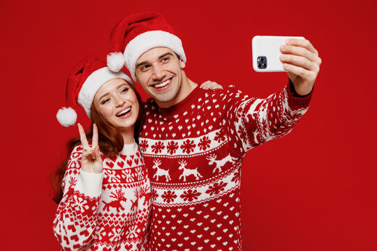 Young couple friends two man woman in sweater hat doing selfie shot on mobile cell phone show v-sign isolated on plain red background. Happy New Year 2022 celebration merry ho x-mas holiday concept.