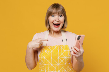 Elderly amazed housekeeper housewife woman 40s in orange apron hold in hand mobile phone search look for recipe do winner gesture isolated plain on yellow background People household lifestyle concept - Powered by Adobe