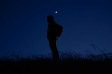 one lonely man tourist with backpack standing among windy field at night with moon shining above...