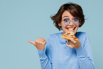 Young woman 20s in casual sweater biting eat slice of pizza point index finger aside on copy space...