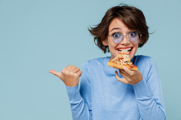 Young woman 20s in casual sweater biting eat slice of pizza point index finger aside on copy space area isolated on plain pastel light blue background studio portrait. People lifestyle food concept.