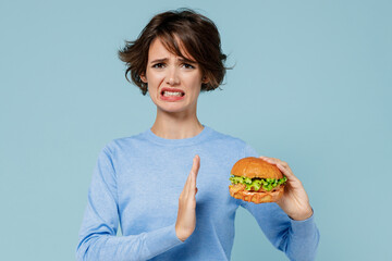 Young sad fun caucasian woman 20s wearing casual sweater hold burger show stop palm gesture reject isolated on plain pastel light blue background studio portrait. People lifestyle junk food concept.